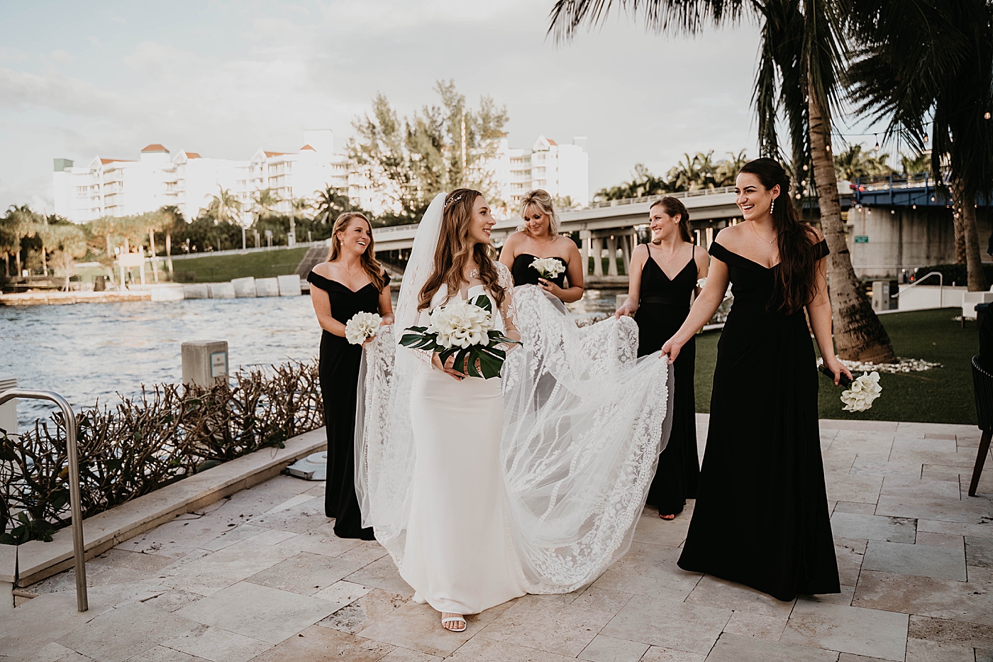 Bride walking with Bridesmaids holding train with palm trees in the background Waterstone Resort and Marina Wedding captured by South Florida Wedding Photographer Krystal Capone Photography