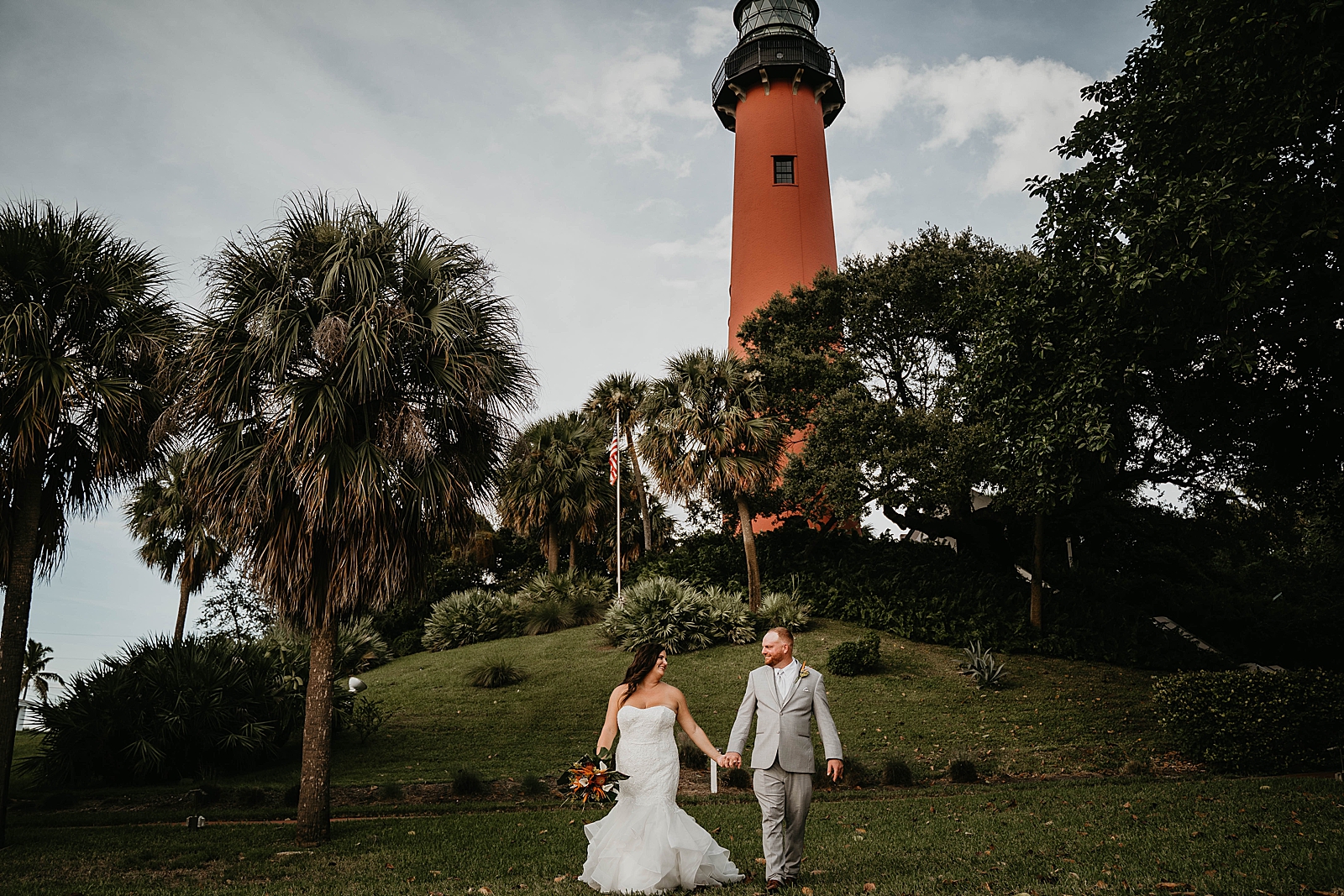 Bride and groom walking down the hill holding hands with palm trees and lighthouse in the background Jupiter Lighthouse Wedding Photography captured by South Florida Wedding Photographer Krystal Capone Photography
