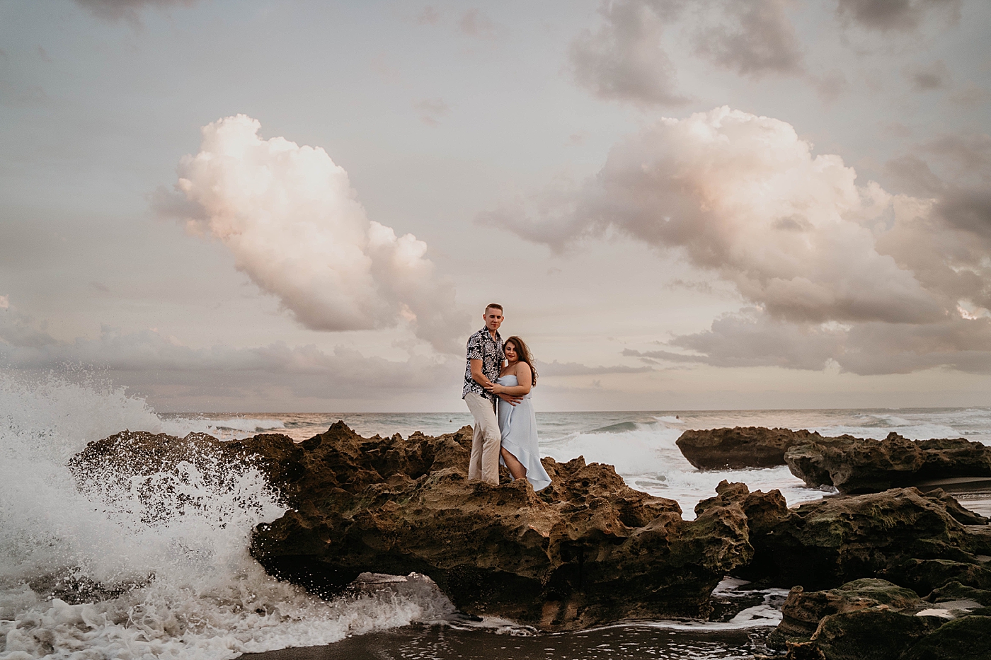Amazing shot of Couple holding each other on top of shoreline rocks with waves coming in South Florida Beach Photography captured by Krystal Capone Photography