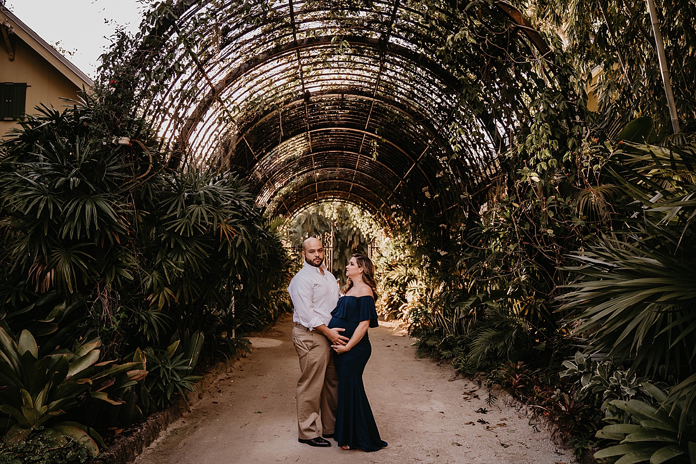 Husband and wife holding bump under green arch way South Florida Maternity Photography captured by South Florida Family Photographer Krystal Capone Photography 
