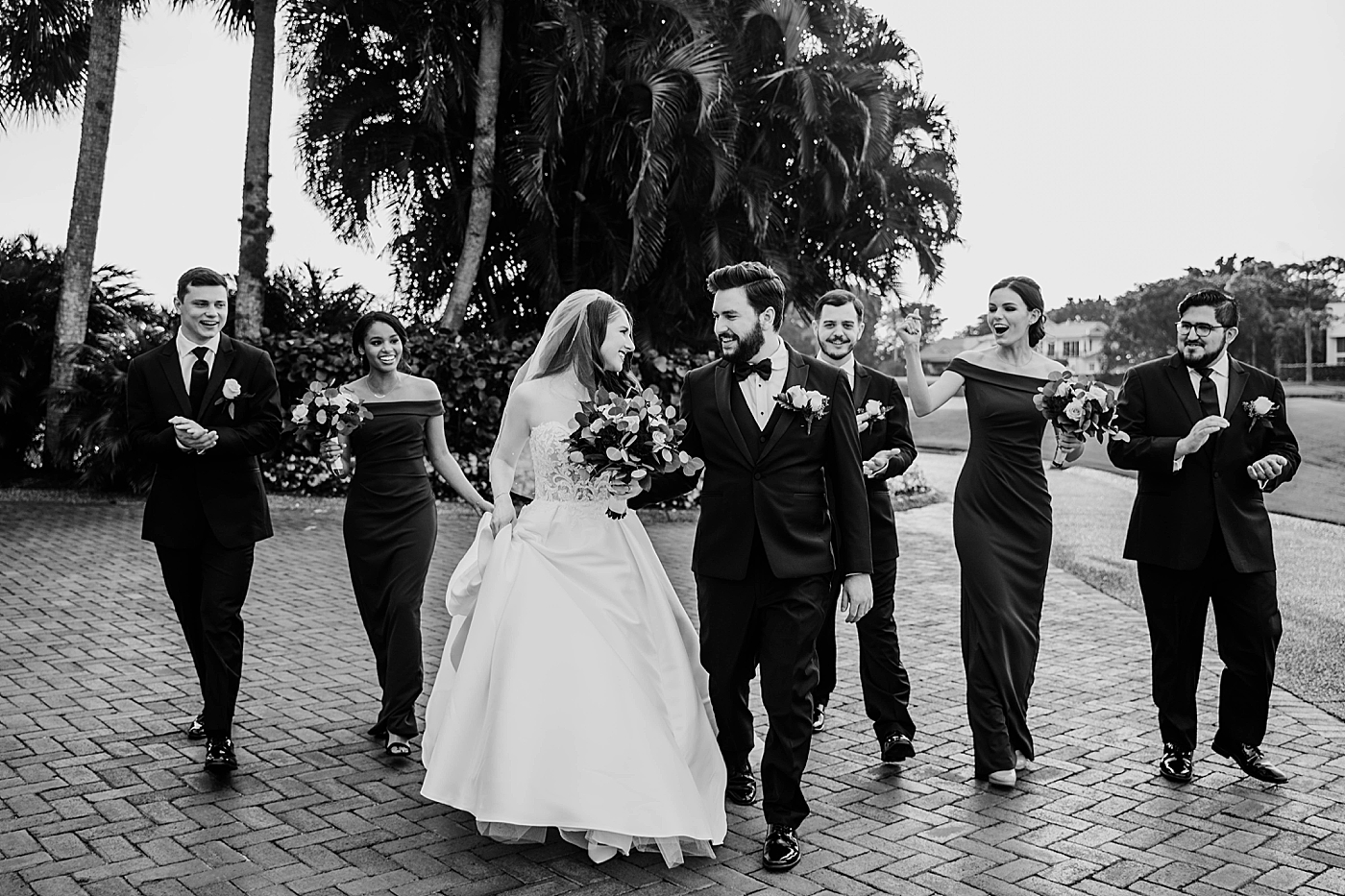 Black and White shot of Bride and Groom walking looking at each other with wedding party following Breakers West Wedding Photography captured by South Florida Wedding Photographer Krystal Capone Photography 