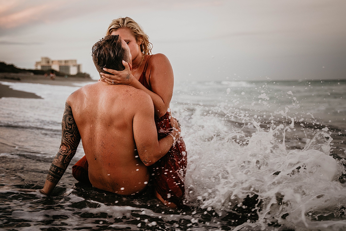 Couple sitting and holding each other and kissing as wave crashes in Jupiter Beach Engagement Photography captured by South Florida Engagement Photographer Krystal Capone Photography 