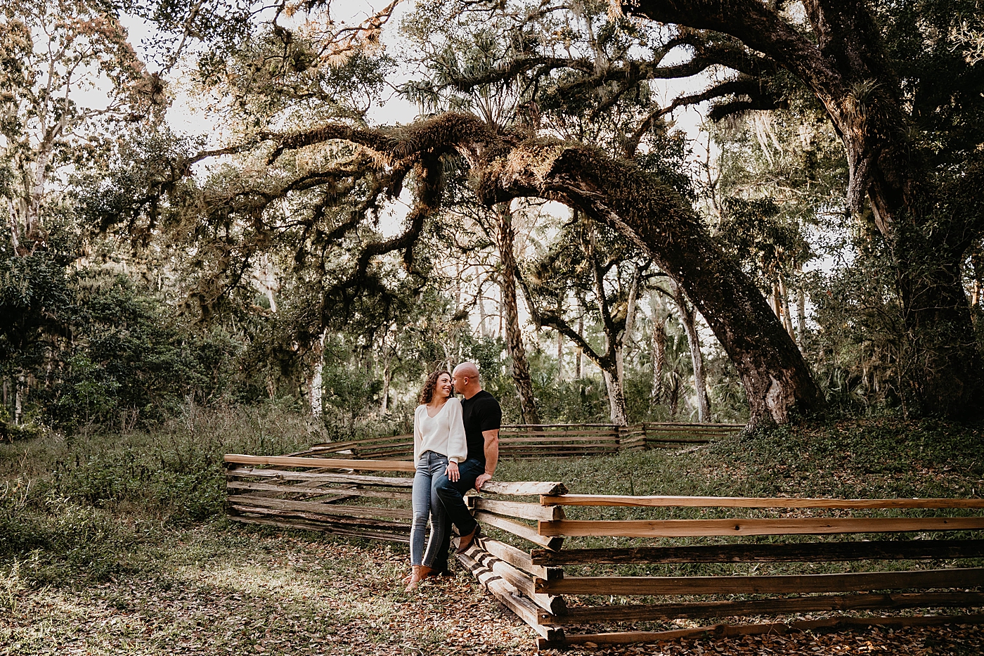 Couple leaning on wood fence with large spanish moss trees Romantic Riverbend Park Engagement Photography captured by South Florida Photographer Krystal Capone Photography