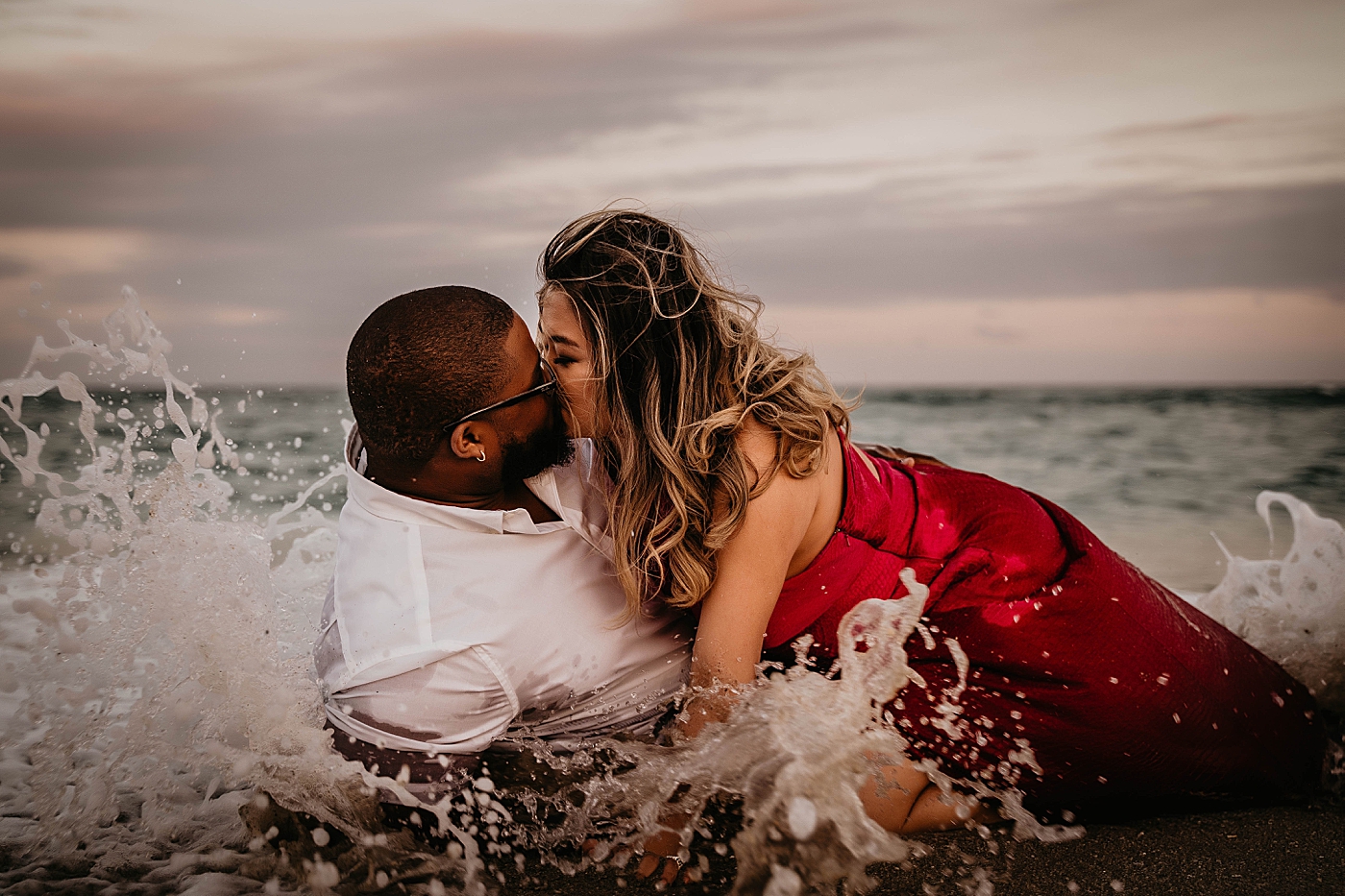 Wife on top of husband kissing in ocean water with waves crashing Palm Beach Anniversary Photography captured by South Florida Family Photographer Krystal Capone Photography