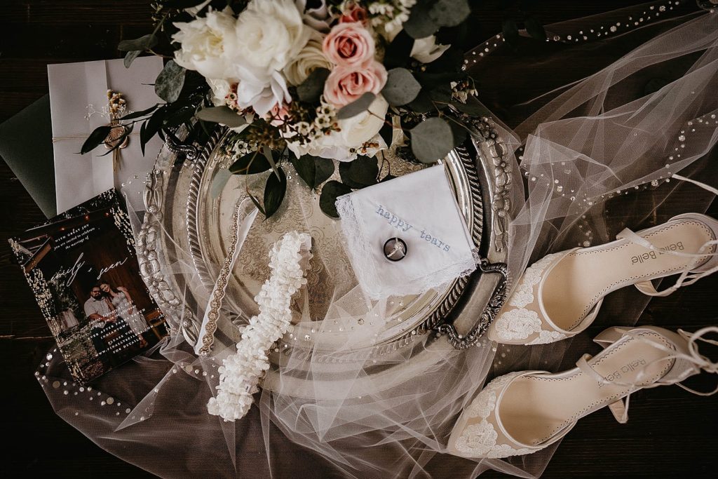 Wedding detail shot of bouquet wedding bands engagement ring and garters on silver platter with white heel and invitation Ever After Farms Wedding Photography captured by South Florida Wedding Photographer Krystal Capone Photography 