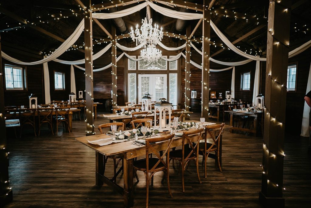 Wide shot of Reception area with rectangular tables with candle lamp centerpieces and bulb lights wrapped around beams Ever After Farms Wedding Photography captured by South Florida Wedding Photographer Krystal Capone Photography 