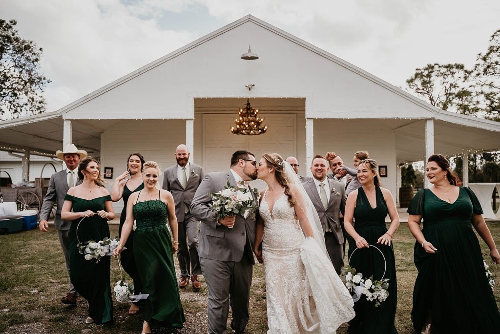 Bride and Groom walking and kissing with Bridal party behind them Ever After Farms Wedding Photography captured by South Florida Wedding Photographer Krystal Capone Photography 