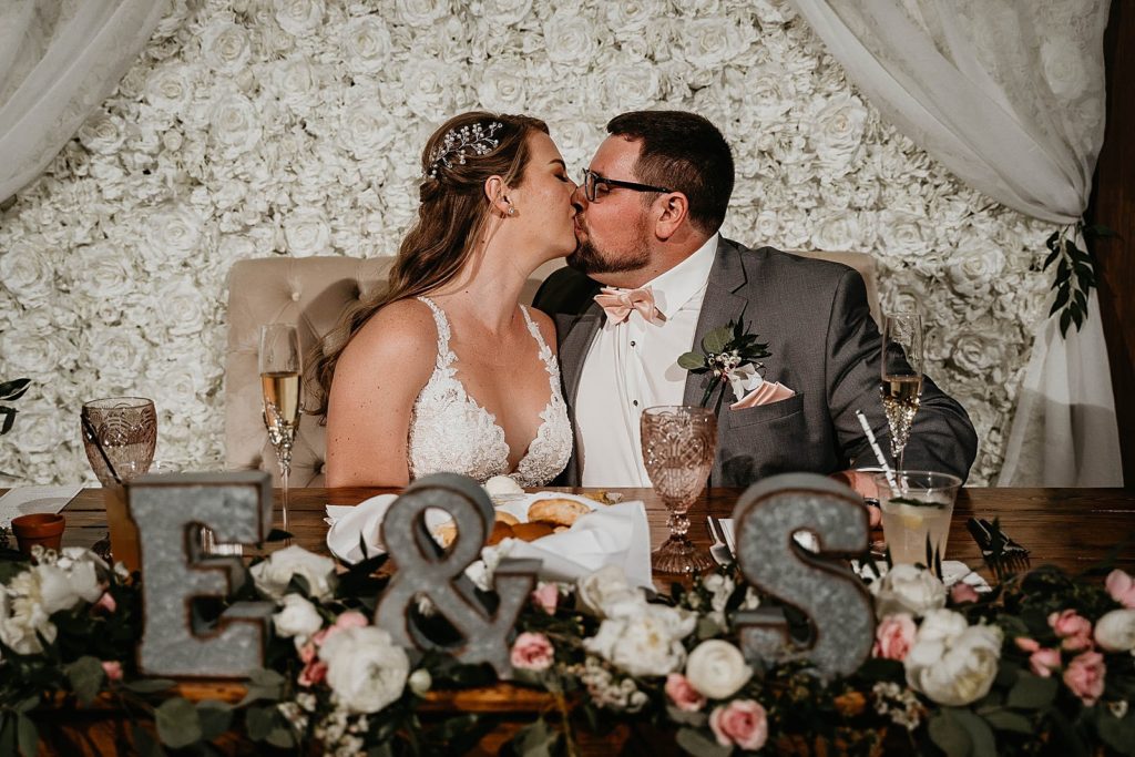 Bride and Groom kissing sweetheart table with white flower wall behind them Ever After Farms Wedding Photography captured by South Florida Wedding Photographer Krystal Capone Photography 