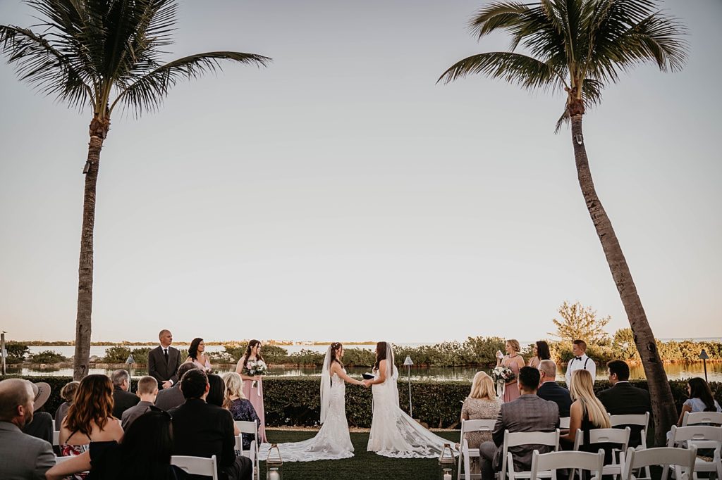 Brides hand in hand wide shot at ceremony next to the water with palm trees Hawks Cay Resort Wedding Photography captured by South Florida Wedding Photographer Krystal Capone Photography