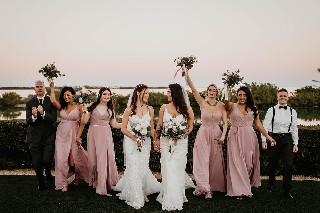 Brides walking together looking at each other with wedding party following and celebrating behind them Hawks Cay Resort Wedding Photography captured by South Florida Wedding Photographer Krystal Capone Photography