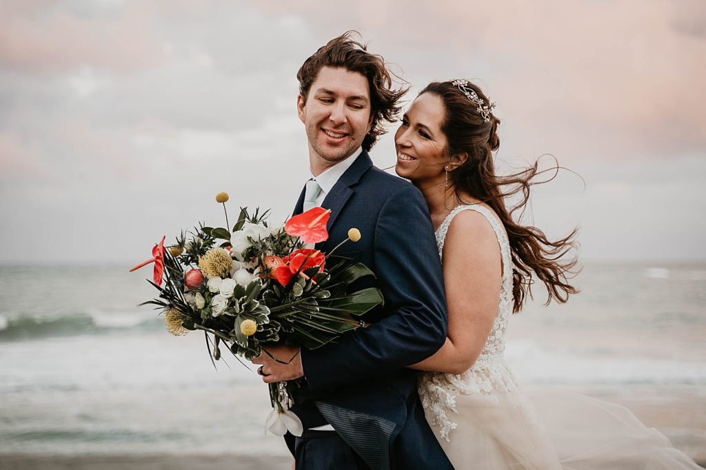 Bride holding Groom from behind with Groom holding tropical bouquet with the ocean behind them Jupiter Beach Resort Wedding Photography captured by South Florida Wedding Photographer Krystal Capone Photography 