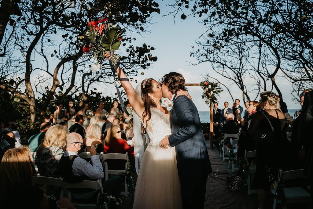 Bride and Groom kissing during Ceremony exit with Bride's extending hand in air with tropical bouquet Jupiter Beach Resort Wedding Photography captured by South Florida Wedding Photographer Krystal Capone Photography 