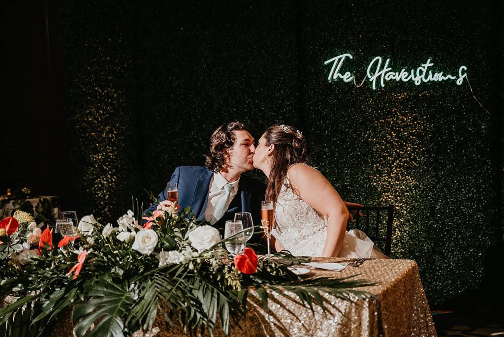 Bride and Groom kissing at sweetheart table with flowers and palm leafs Jupiter Beach Resort Wedding Photography captured by South Florida Wedding Photographer Krystal Capone Photography 