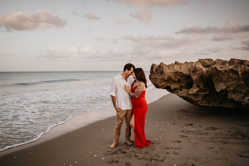 Couple holding each other close on the sand by beach rock and calm ocean water Palm Beach Engagement Photography captured by South Florida Engagement Photographer Krystal Capone Photography 