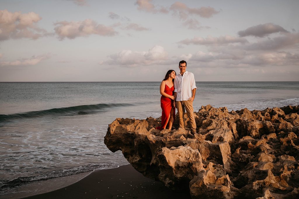 Couple standing together on drift rock on the beach with calm ocean water Palm Beach Engagement Photography captured by South Florida Engagement Photographer Krystal Capone Photography 