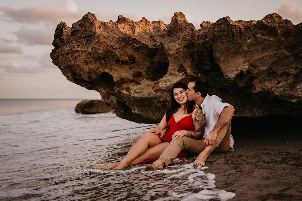 Couple sitting by beach rock with ocean water coming in with man kissing woman on the cheek Palm Beach Engagement Photography captured by South Florida Engagement Photographer Krystal Capone Photography 