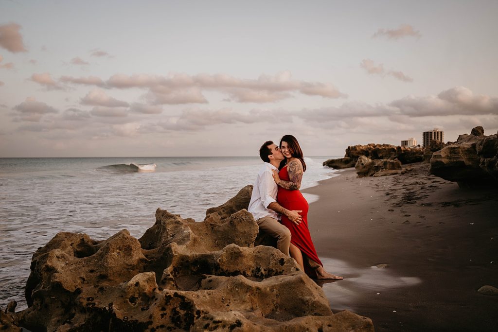Man sits on drift rock on the beach holding woman close and kissing her Palm Beach Engagement Photography captured by South Florida Engagement Photographer Krystal Capone Photography 