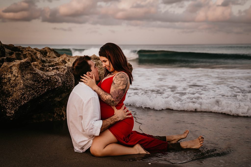 Man sitting on wet sand by beach rock with lady saddling on his lap as they look into each others eyes Palm Beach Engagement Photography captured by South Florida Engagement Photographer Krystal Capone Photography 