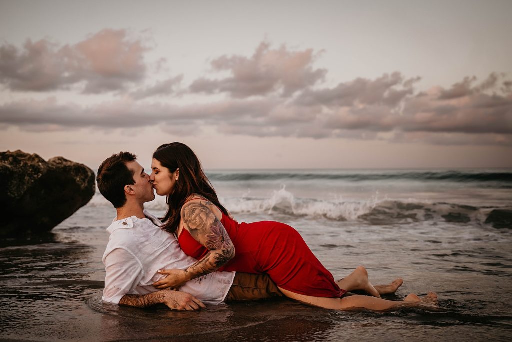 Lady on top of man on the beach kissing him as waves come in Palm Beach Engagement Photography captured by South Florida Engagement Photographer Krystal Capone Photography 