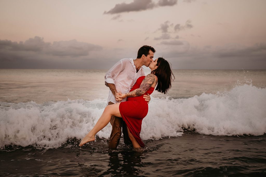 Couple kissing on the beach with a wave about to hit them Palm Beach Engagement Photography captured by South Florida Engagement Photographer Krystal Capone Photography 