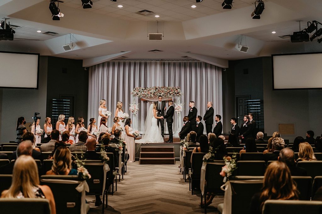 Wide shot of Wedding attendees Bridal Party Bride and Groom holding hands as they listen to the Homily Pelican Club Wedding Photography captured by South Florida Wedding Photographer Krystal Capone Photography