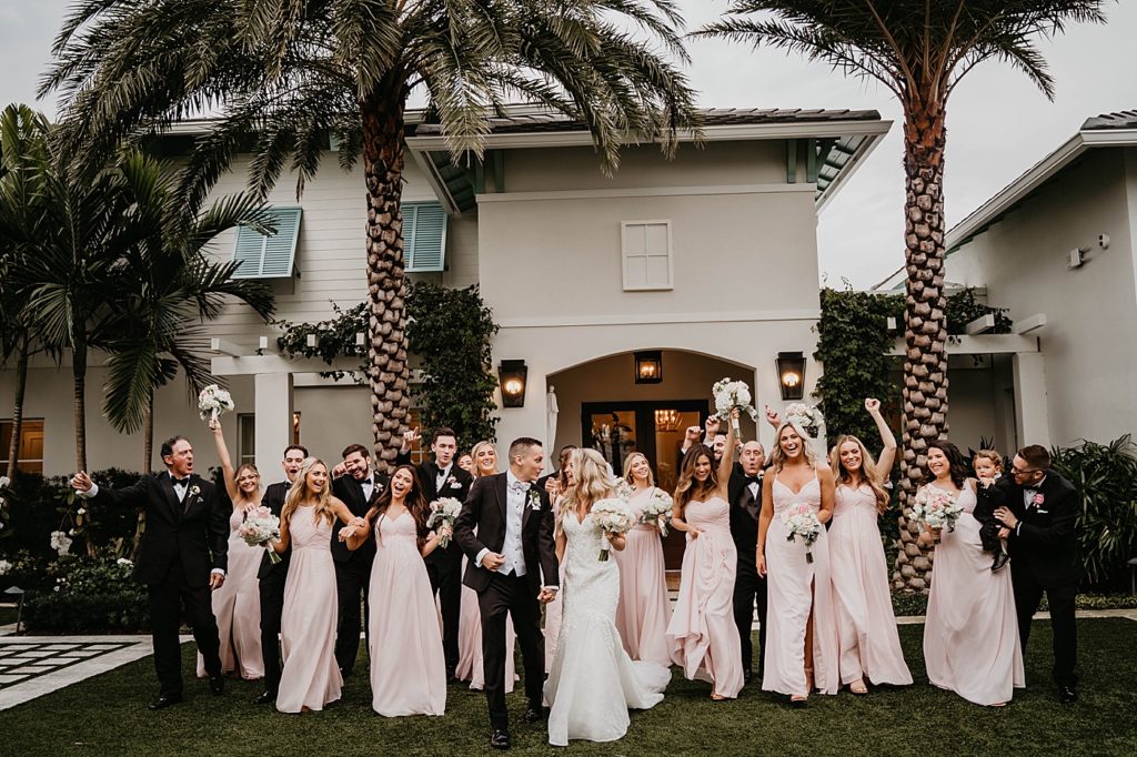 Bride and Groom holding hands behind and looking at each other with Wedding party celebrating behind them Pelican Club Wedding Photography captured by South Florida Wedding Photographer Krystal Capone Photography