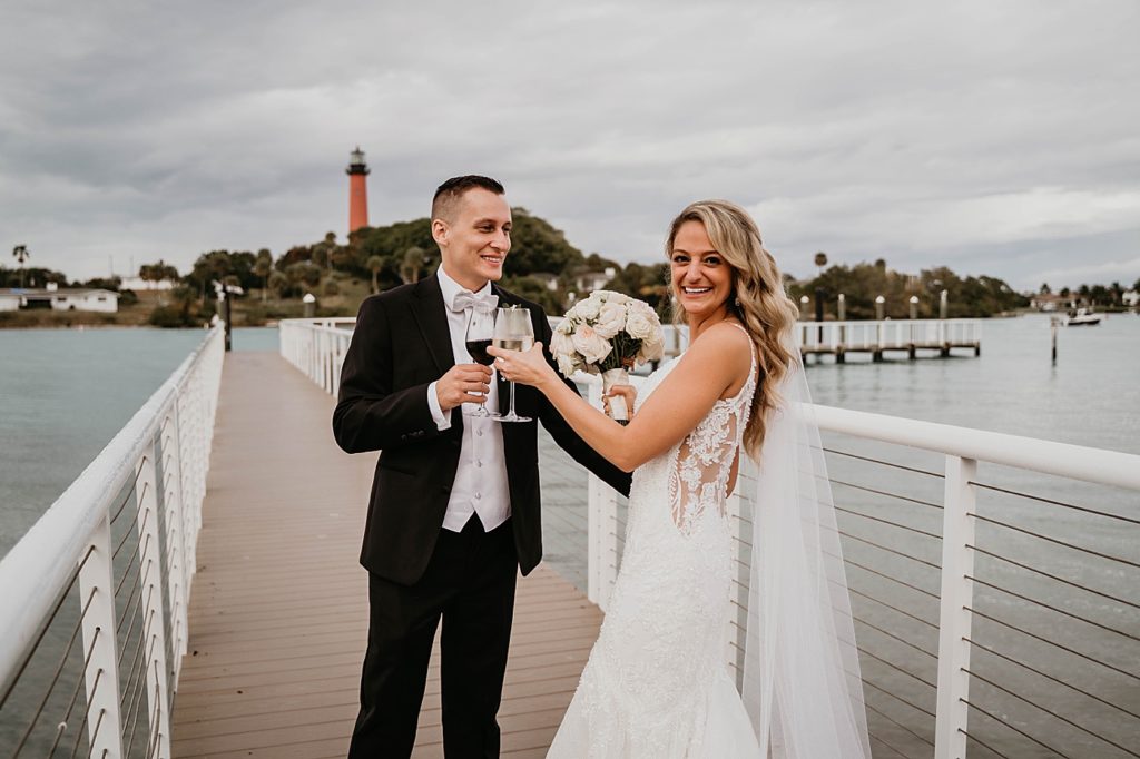 Bride and Groom cheering each other with red and white wine on bridge with lighthouse in the distance Pelican Club Wedding Photography captured by South Florida Wedding Photographer Krystal Capone Photography