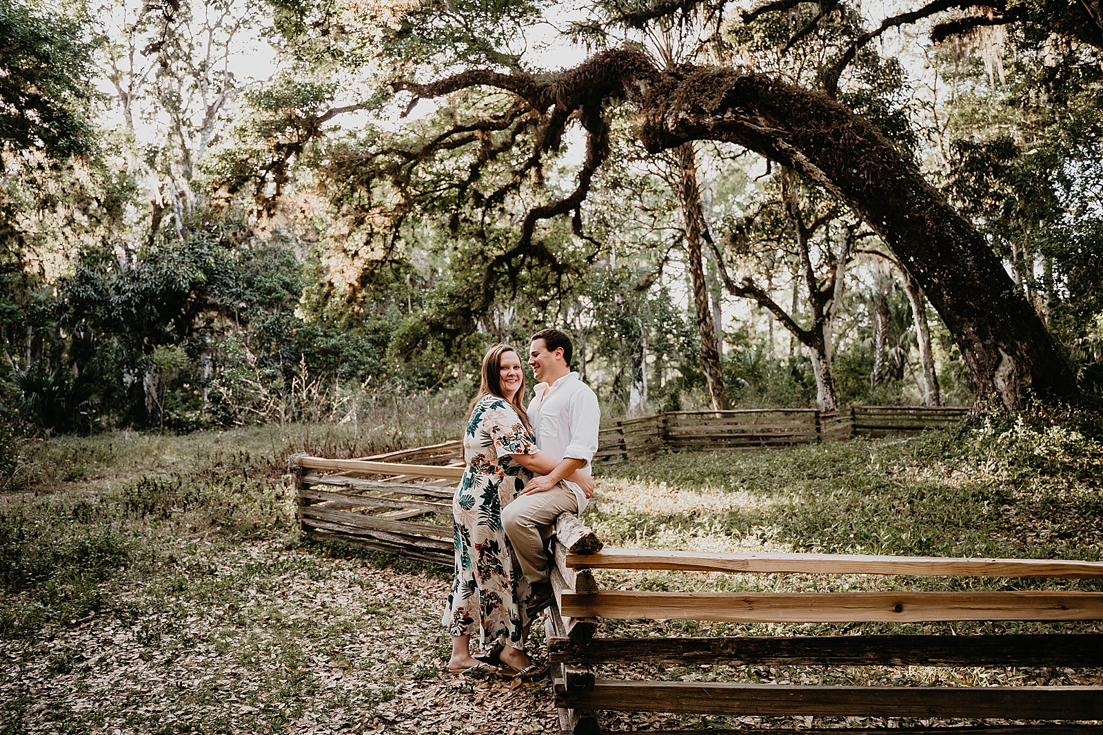 Woman holds man who is sitting on rustic wooden fence in forest Riverbend Park Engagement Photography captured by South Florida Engagement Photographer Krystal Capone Photography 