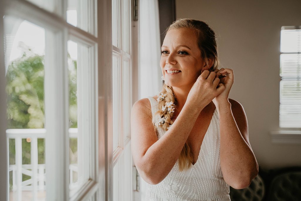 Bride getting ready putting on earrings by the window The Lake House Fort Pierce Wedding Photography captured by South Florida Wedding Photographer Krystal Capone Photography 