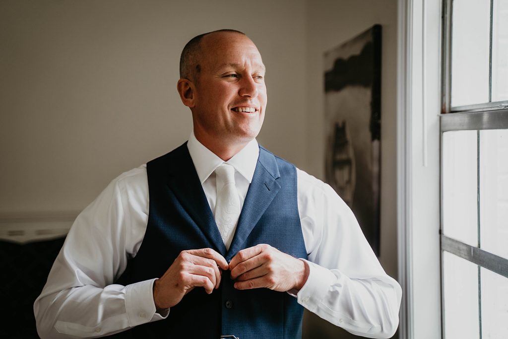 Groom getting ready buttoning up vest by the window The Lake House Fort Pierce Wedding Photography captured by South Florida Wedding Photographer Krystal Capone Photography 