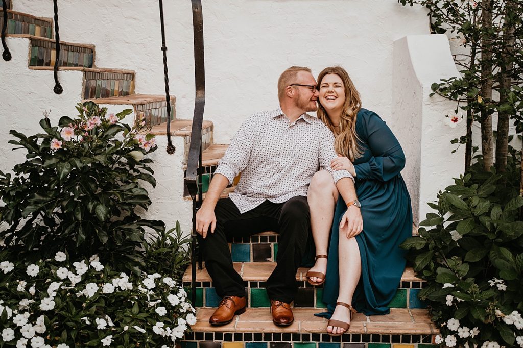 Couple sitting together nuzzling on staircase with greenery and flowers Worth Ave Palm Beach Engagement Photography captured by South Florida Engagement Photographer Krystal Capone Photography