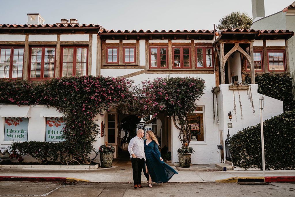 Wide shot of couple holding each other by the side in front of Piccolo Mondo with lots of greenery and pink flowers Worth Ave Palm Beach Engagement Photography captured by South Florida Engagement Photographer Krystal Capone Photography