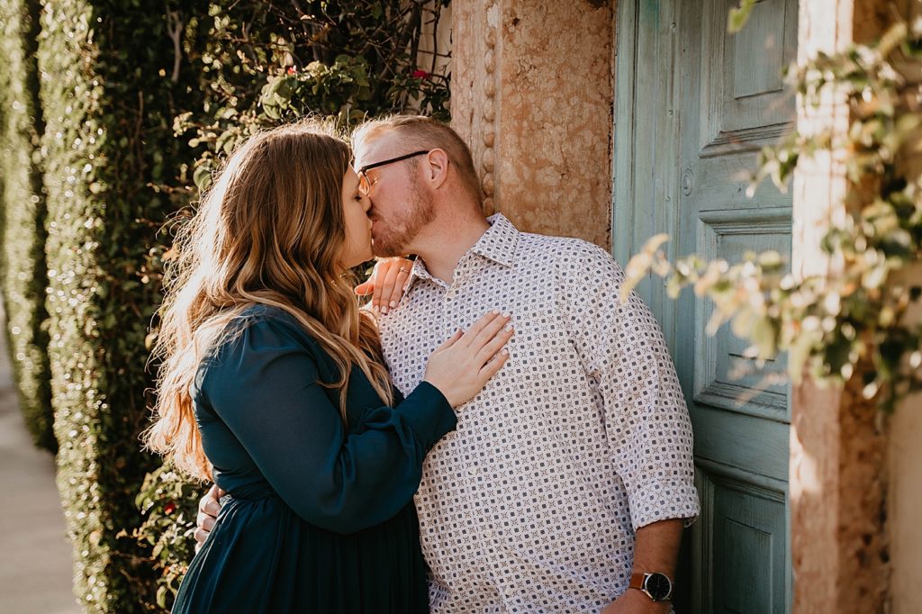 Couple kissing holding each other in front of turquoise door and hedges Worth Ave Palm Beach Engagement Photography captured by South Florida Engagement Photographer Krystal Capone Photography