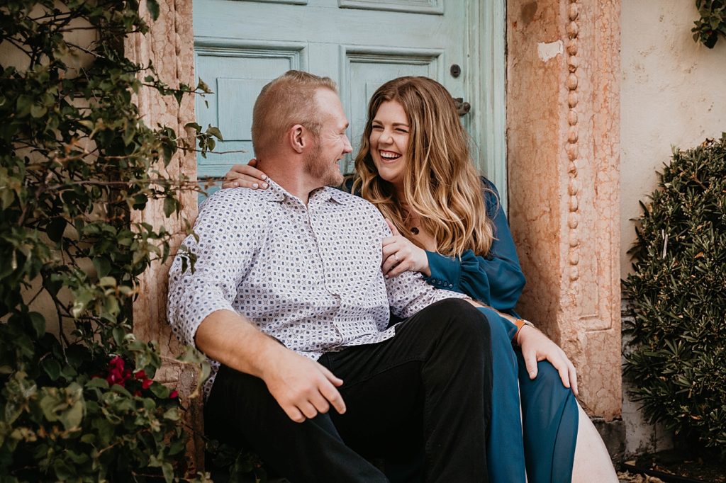 Couple sitting down in front of turquoise blue door smiling and looking at each other Worth Ave Palm Beach Engagement Photography captured by South Florida Engagement Photographer Krystal Capone Photography