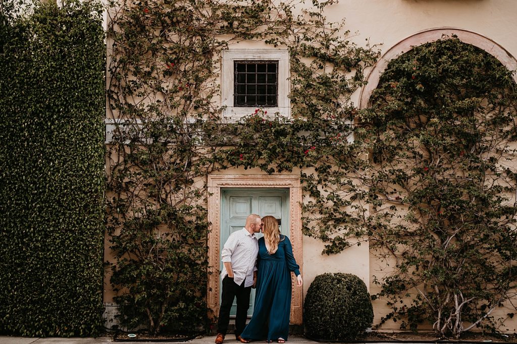 Couple kiss in front of house with greenery growing on all over Worth Ave Palm Beach Engagement Photography captured by South Florida Engagement Photographer Krystal Capone Photography