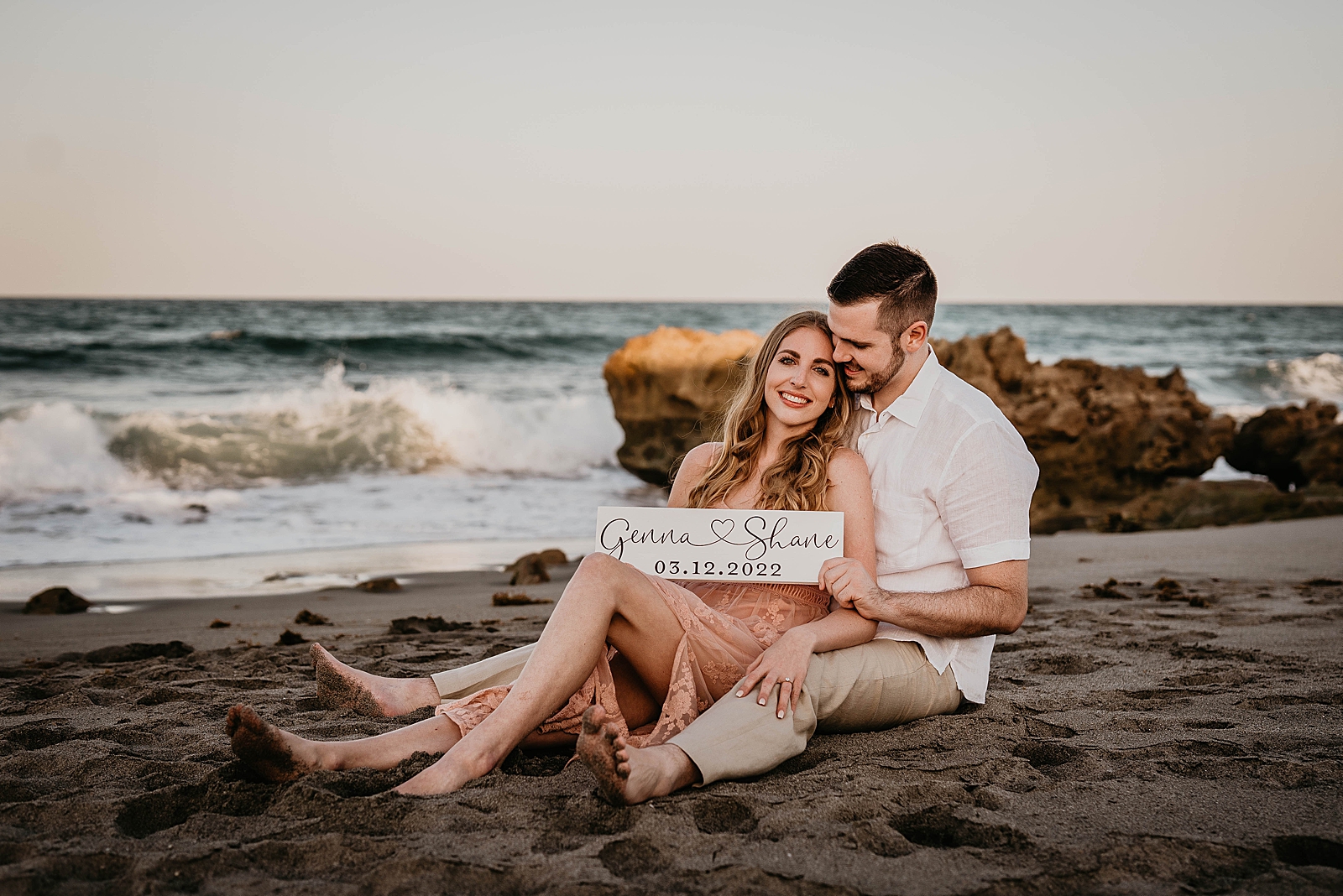 Couple holding sign for save the date by the ocean sitting on the beach