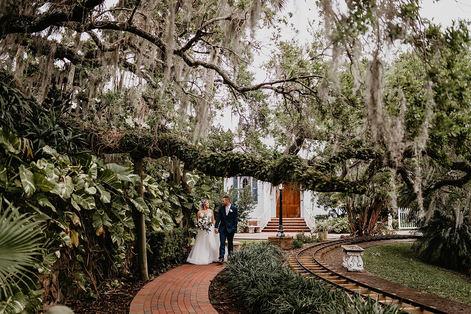 Couple holding hands and walking on brick path under large tree branch with Spanish moss