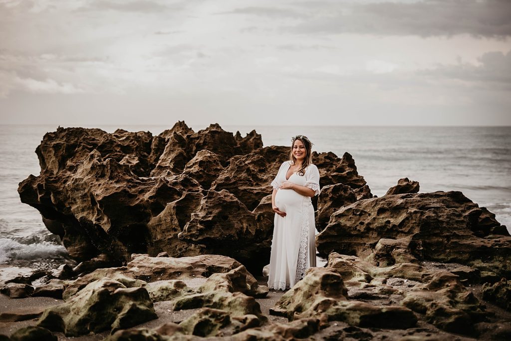 Pregnant woman standing on the beach by the rocks with calm ocean behind her