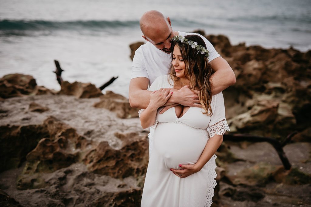 Husband holding pregnant wife with drift rock and waves behind them